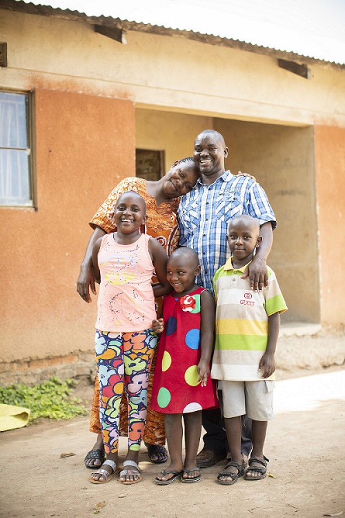 Middle aged man in Uganda stands smiling with his wife and three children.