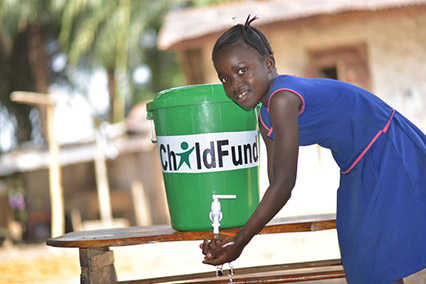 A 10-year-old girl in Sierra Leone washes her hands with water from a green bucket labeled ChildFund.