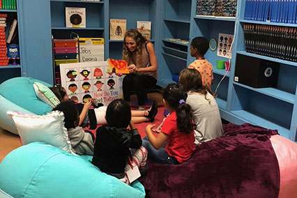 A woman reads a story to a group of children sitting in bean bags in a brightly colored classroom corner in the United States.