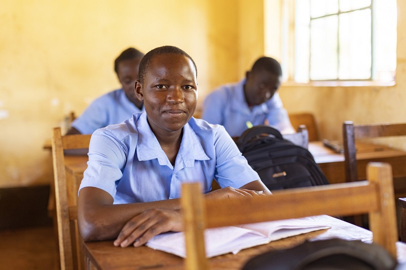 A teen girl in Kenya sits at a desk, slightly smiling.