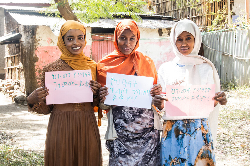 Three young women in Ethiopia hold up signs in Amharic, smiling at the camera.