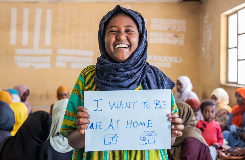 Girl in Ethiopia holds up a sign that reads, "I want to be safe at home," smiling at the camera.