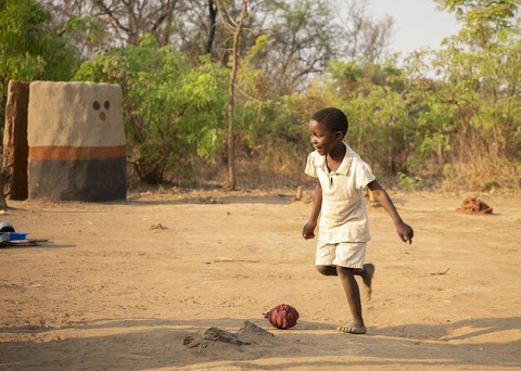 A boy plays soccer with a red homemade ball outside in Zambia.
