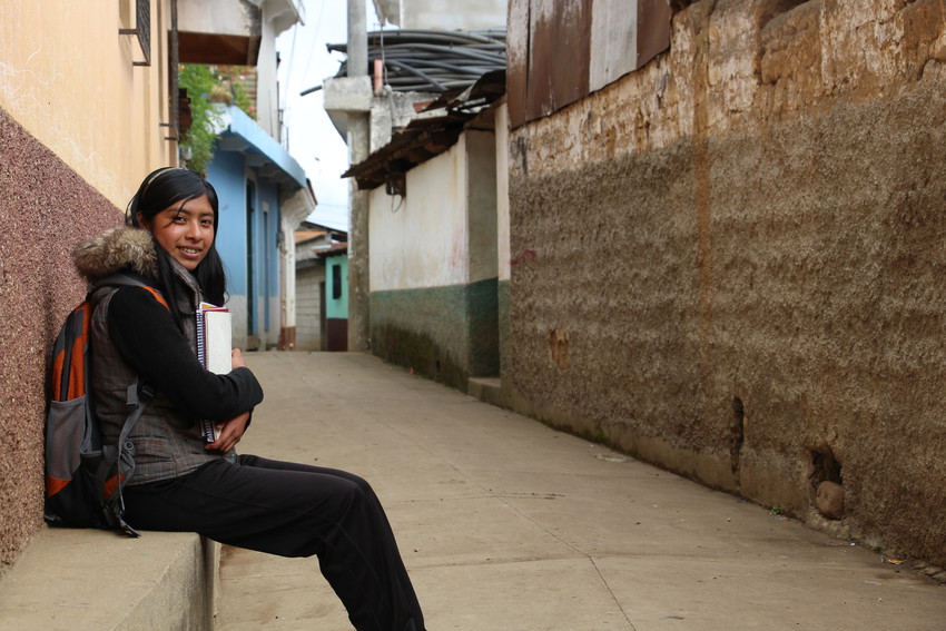 Schoolgirl in Guatemala sits outside in a empty street wearing backpack and holding books, smiling.