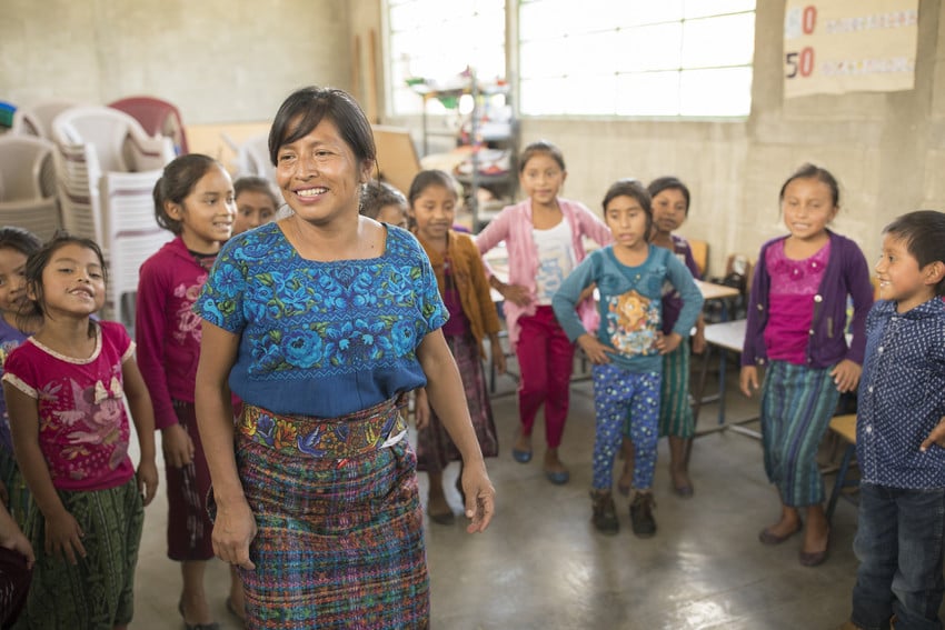 A teacher in a blue traditional Guatemalan dress stands in room full of elementary students at school, smiling.