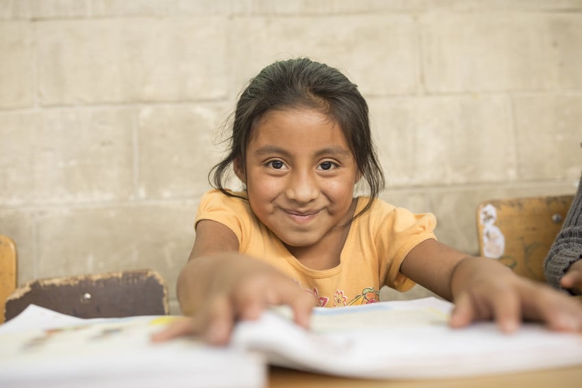 Young girl sits at a desk at school in Guatemala, holding papers, smiling.