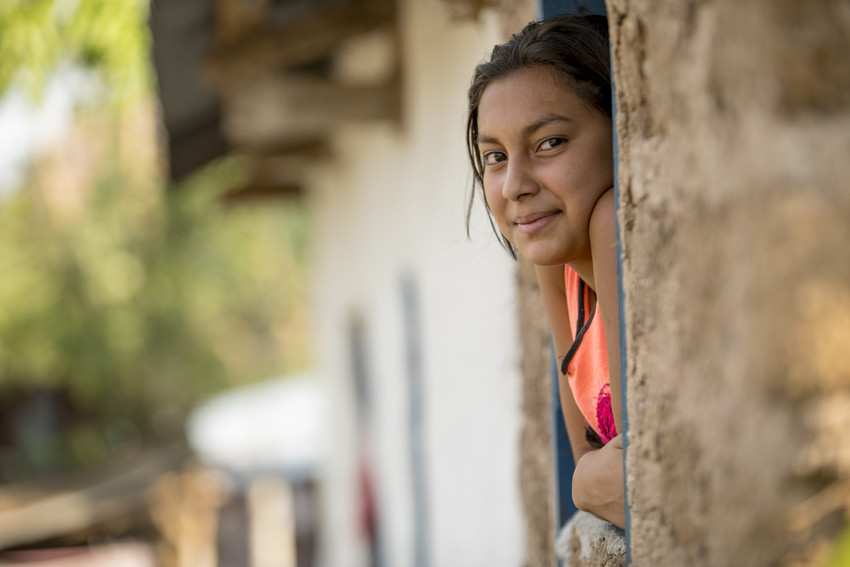 Teenage girl in Santa Barbara Department, Honduras, leaning out of window with arms crossed looking at camera, smiling.