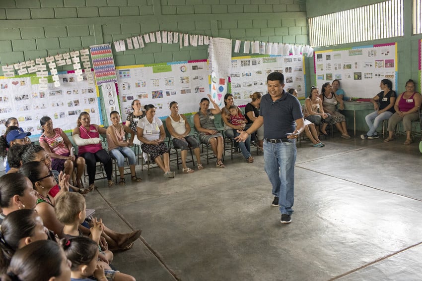 Male teacher at school in Guatemala stands in the middle of a classroom full of parents, talking and gesturing with his hands.